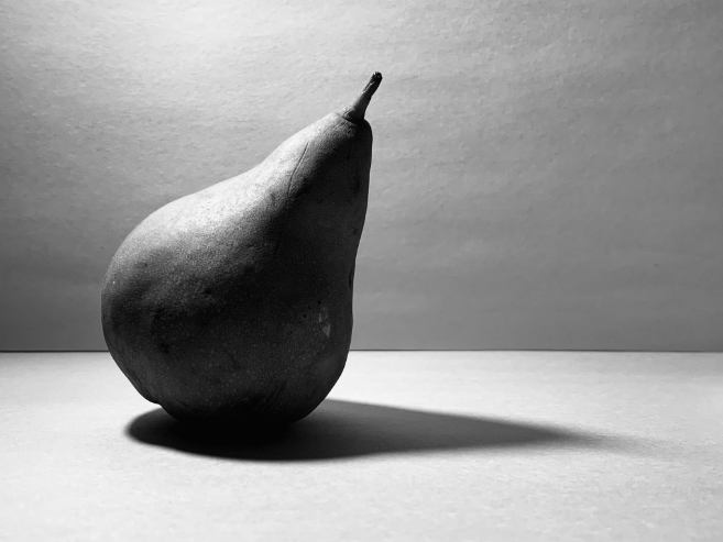 Photograph of a single pear placed on a flat surface with a plain background, captured in black and white. Strong lighting creates a distinct shadow extending to the right, emphasizing the pear's texture and shape.
