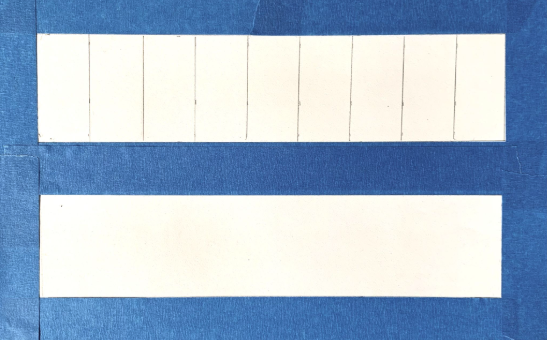 Photograph of two white rectangular strips framed by blue tape, with the top strip divided into ten equal sections by vertical lines and the bottom strip left blank. The layout suggests a comparison purpose, highlighting the segmented versus unsegmented design.