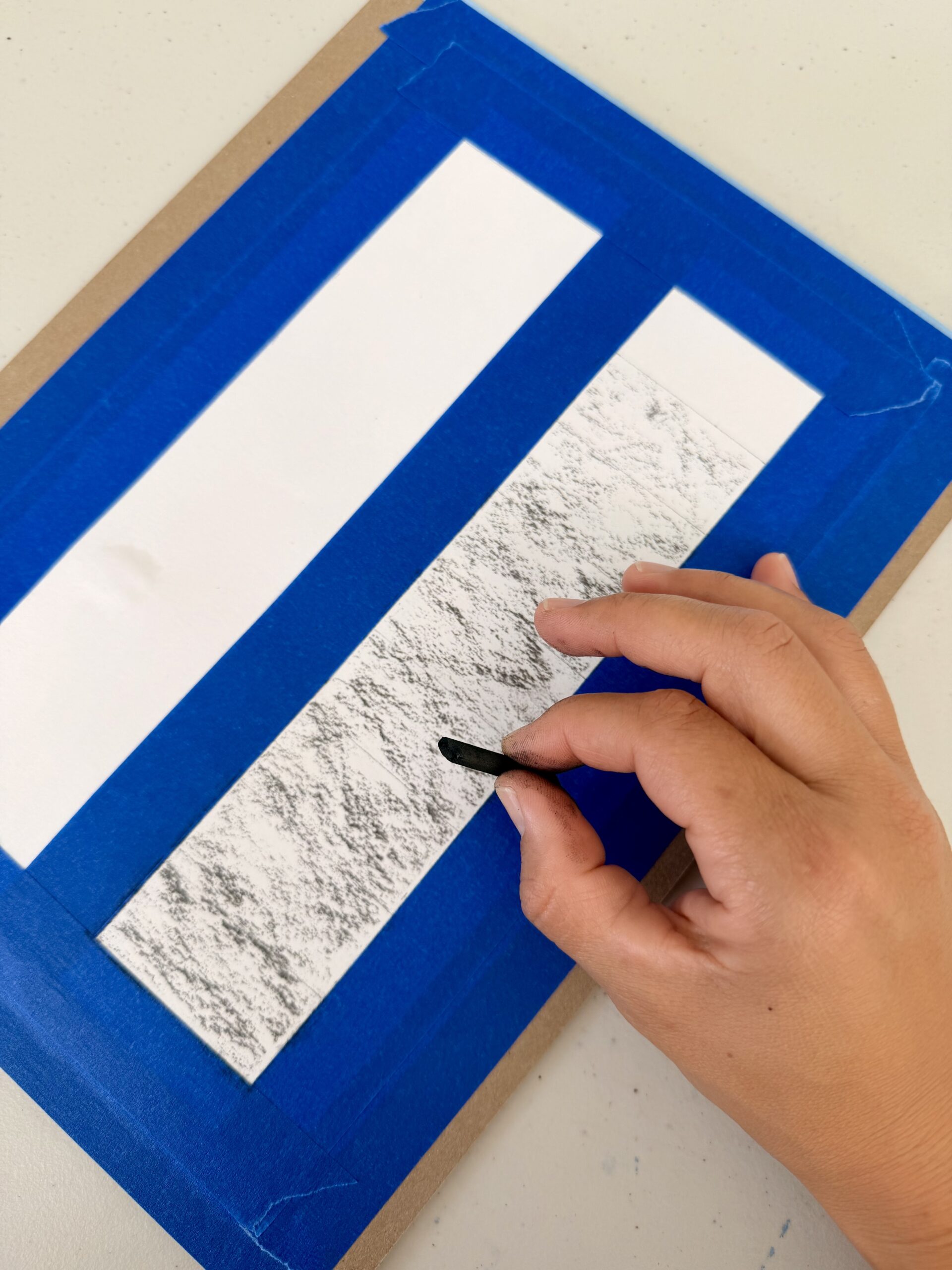 Photograph of a hand using a black vine charcoal to create a textured rubbing on a white strip of paper taped to a surface with blue painter's tape. The rubbing reveals a detailed, rough pattern underneath, contrasting with a blank white strip beside it.