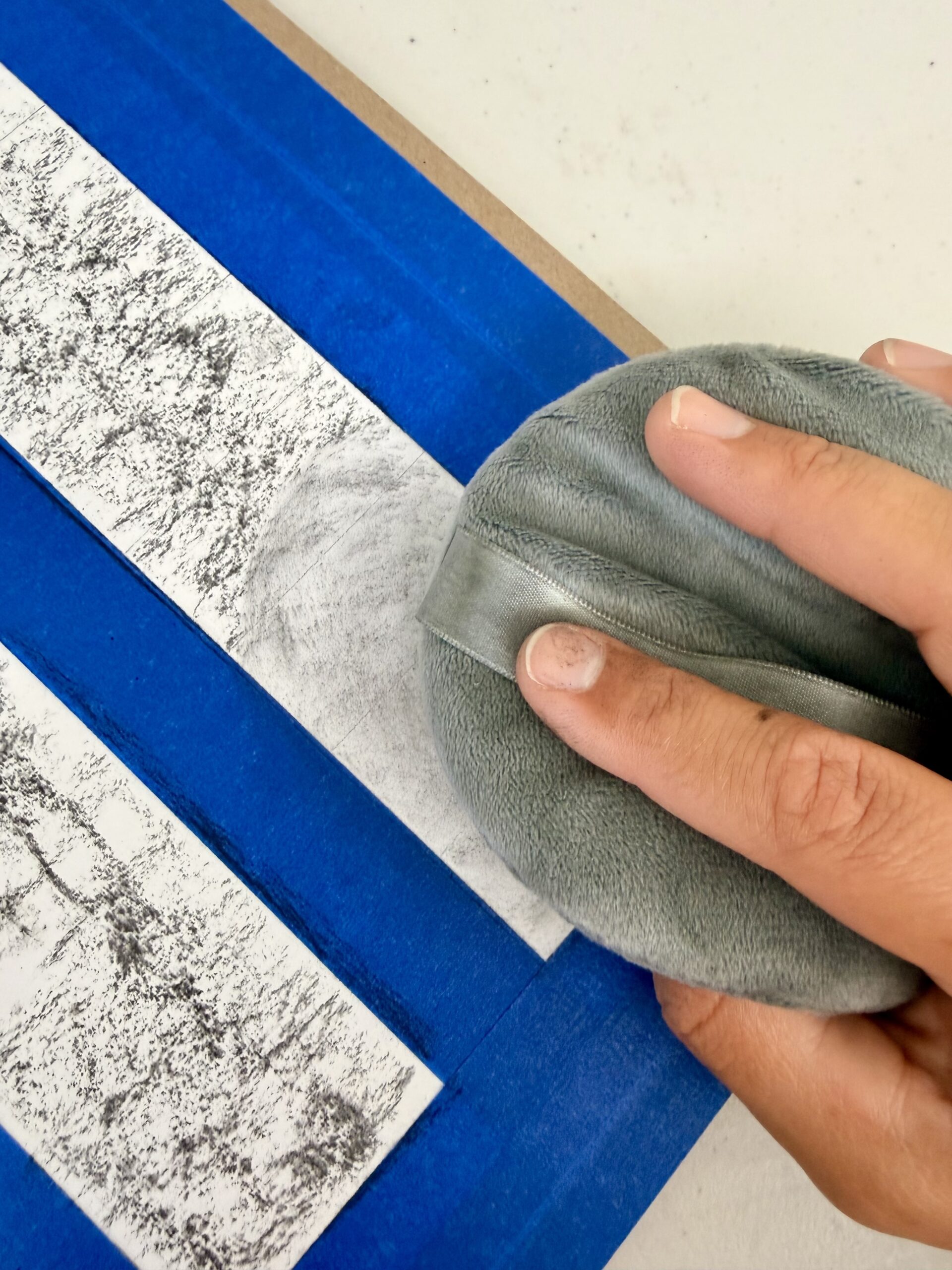 Photograph showing a close-up of a hand using a powder puff on a white surface with black textured patterns, bordered by blue painter's tape. The scene highlights toning process using vine charcoal on drawing paper.