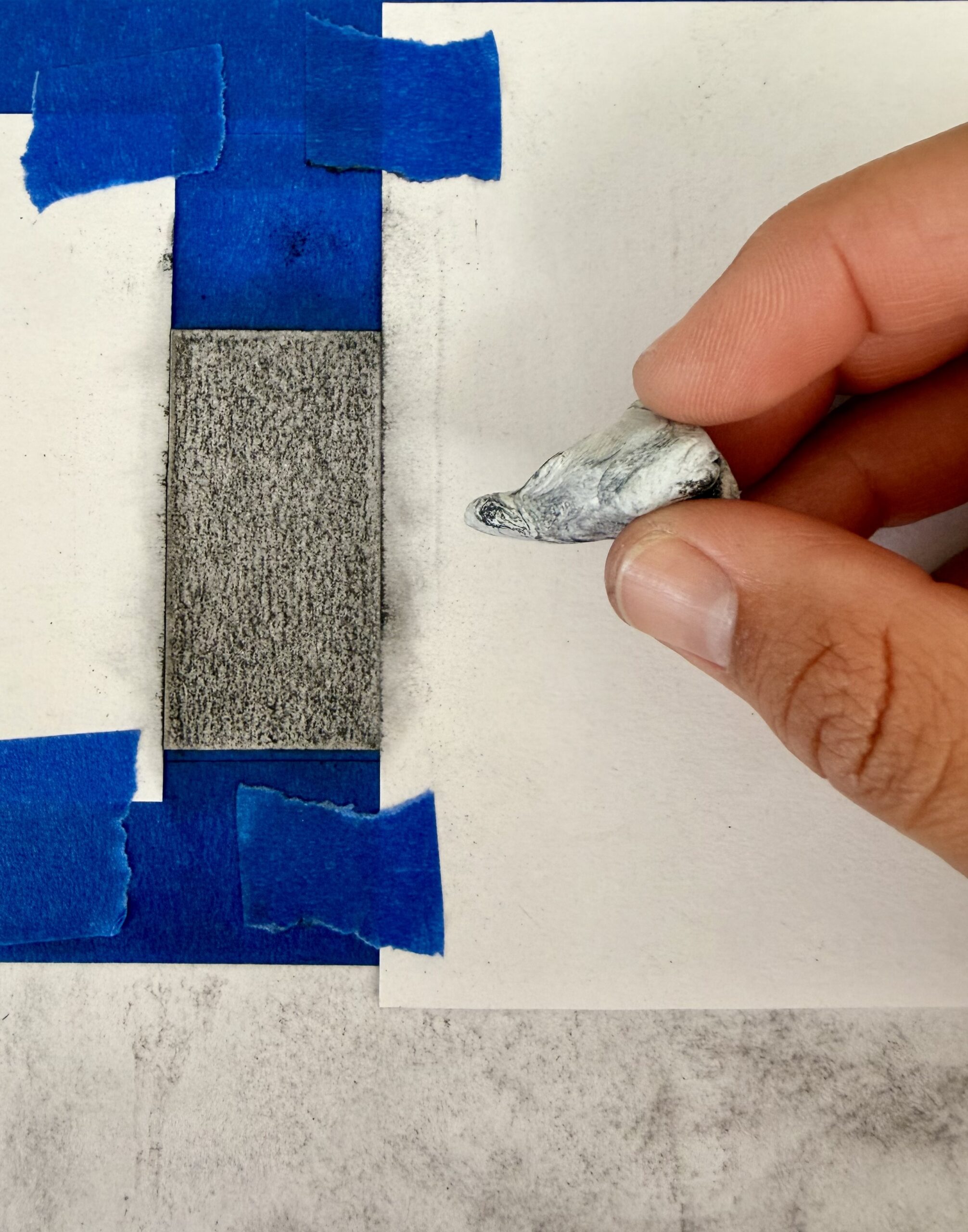 Photograph showing a hand holding a kneaded eraser above a rectangular area shaded with pencil, bordered by blue painter's tape on white paper. The setup suggests a drawing technique demonstration.