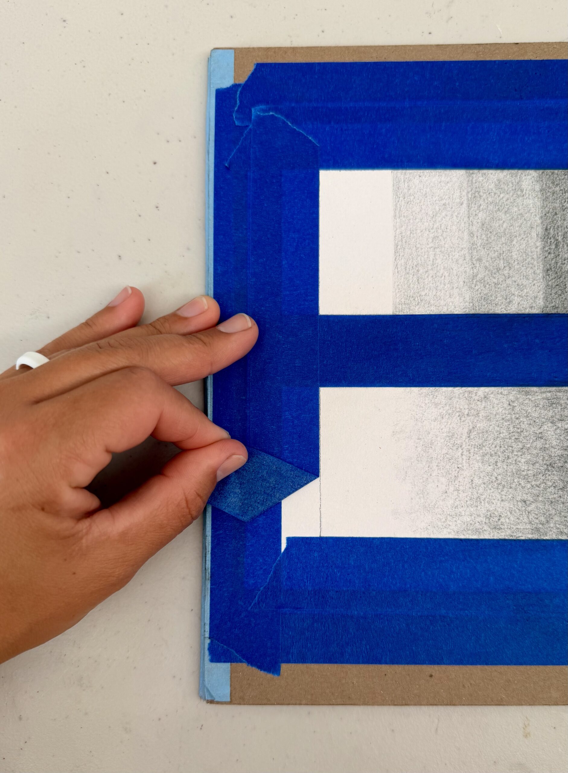 Photograph showing a hand peeling blue painter's tape from a drawing board with a grayscale gradient artwork. The tape frames the artwork in a rectangular shape, highlighting careful removal to reveal clean edges around the shaded gradient.