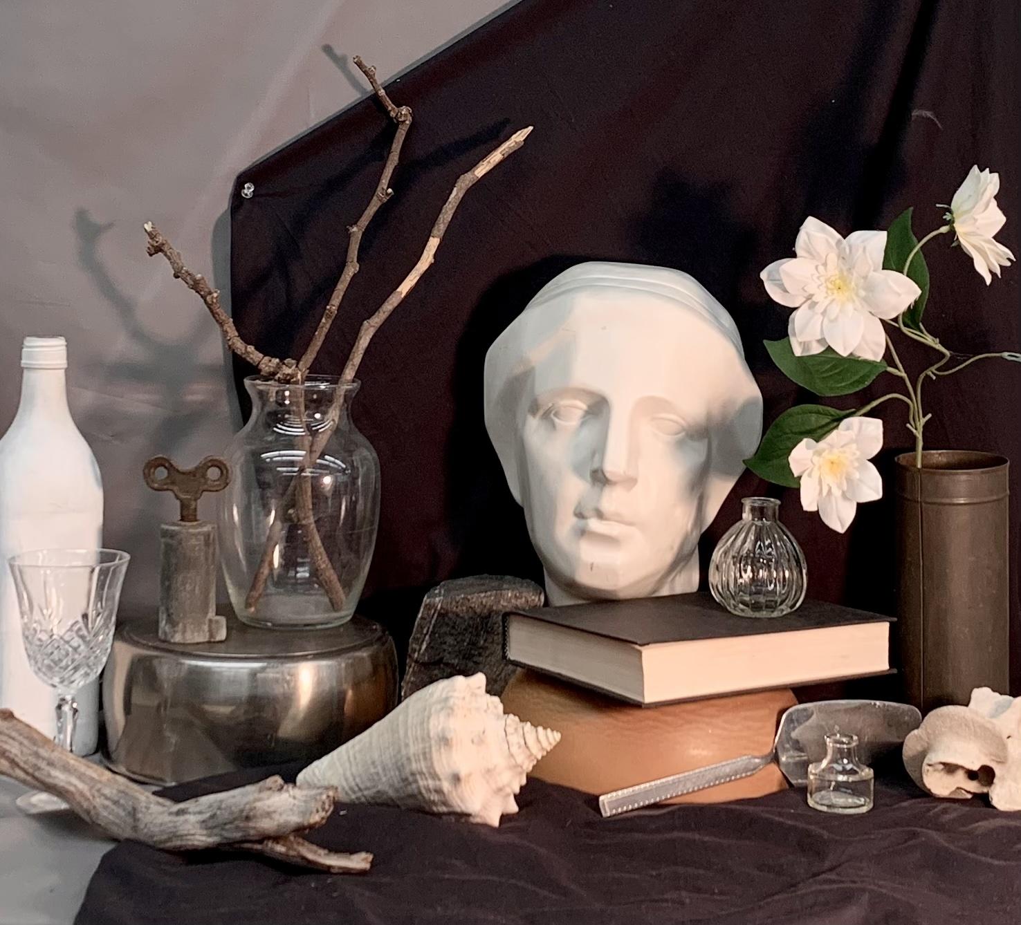 Still life photograph featuring a white classical bust, books, a glass vase with bare branches, and white flowers arranged on a dark cloth backdrop. Additional elements include a large seashell, a glass bottle, a metal key, and a skull, creating a contrast between natural and man-made objects.