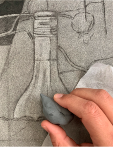 A photograph showing a close-up of a hand using a kneaded eraser to create highlights on a detailed pencil drawing of a bottle and fruit. The drawing features intricate shading and contour lines, with the eraser shaping light areas to enhance depth and realism.