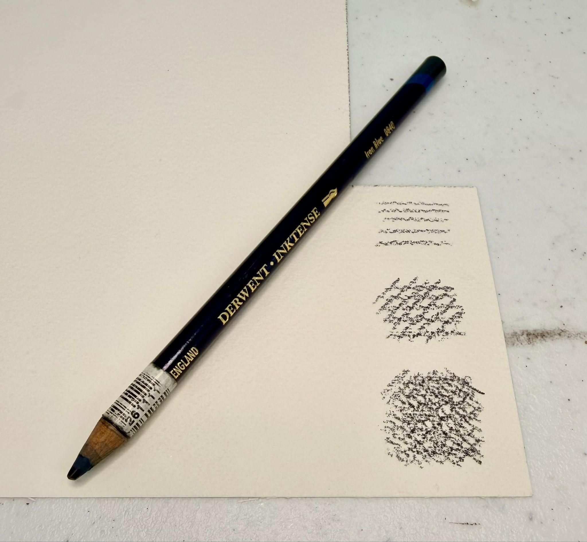 Photograph of a black Derwent Inktense pencil resting on a cream-colored paper with three clusters of markings in varying densities. Pencil features gold lettering and a white barcode sticker near the sharpened tip.