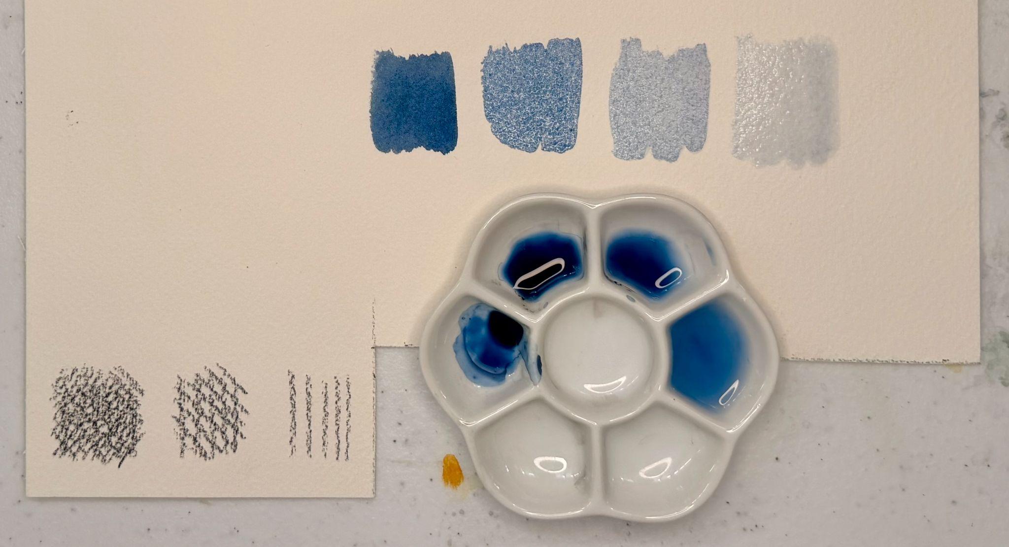 Photograph of a watercolor paint palette with blue paint in several wells, accompanied by two sheets of paper displaying different brushstroke and texture samples in shades of blue. The setup highlights experimentation with paint dilution and brush techniques, showing gradients from dark to light blue and various line and dot patterns.