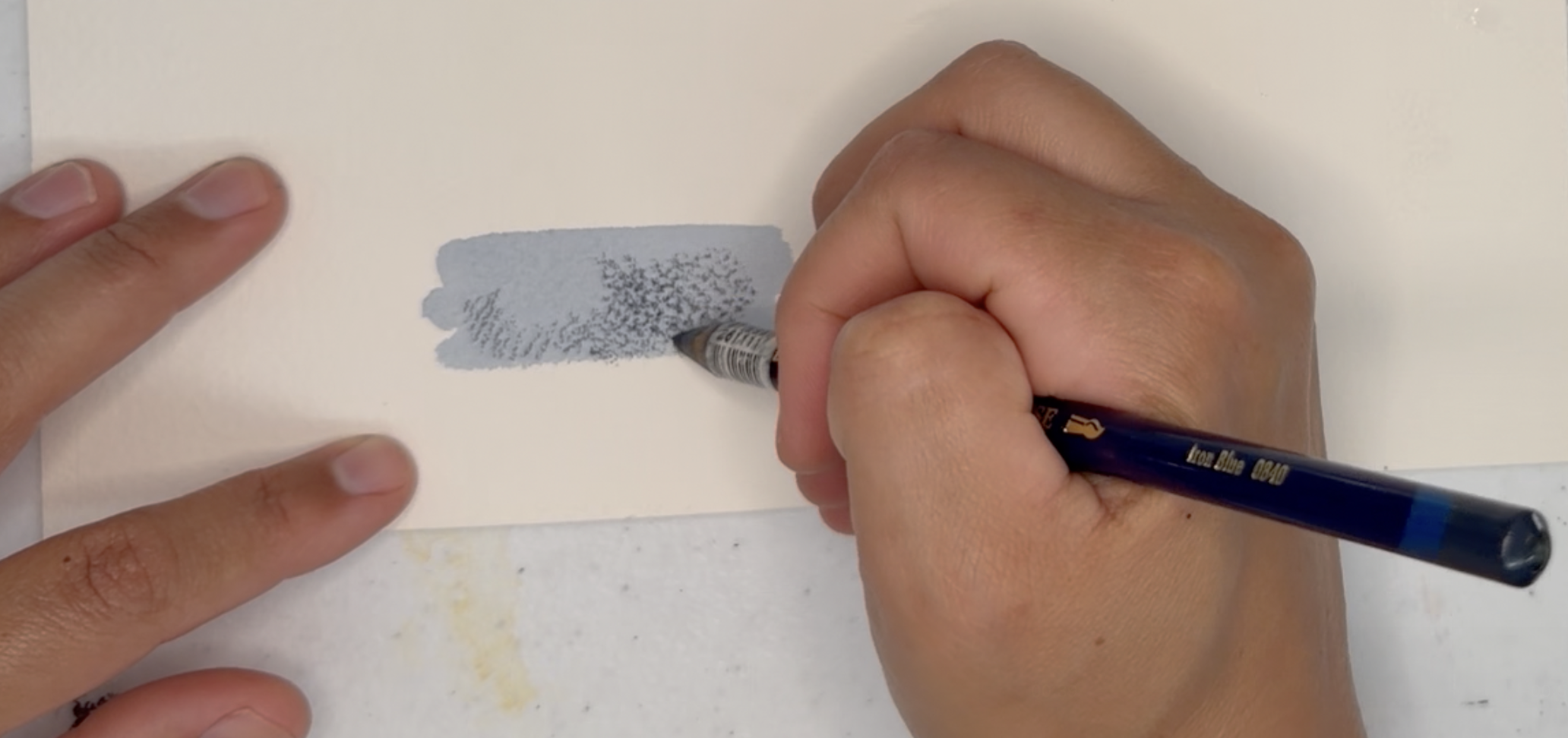 Photograph showing a close-up of a person shading a small rectangular area on white paper using a pencil. The shaded area transitions from light to dark with visible pencil strokes, demonstrating a shading technique.