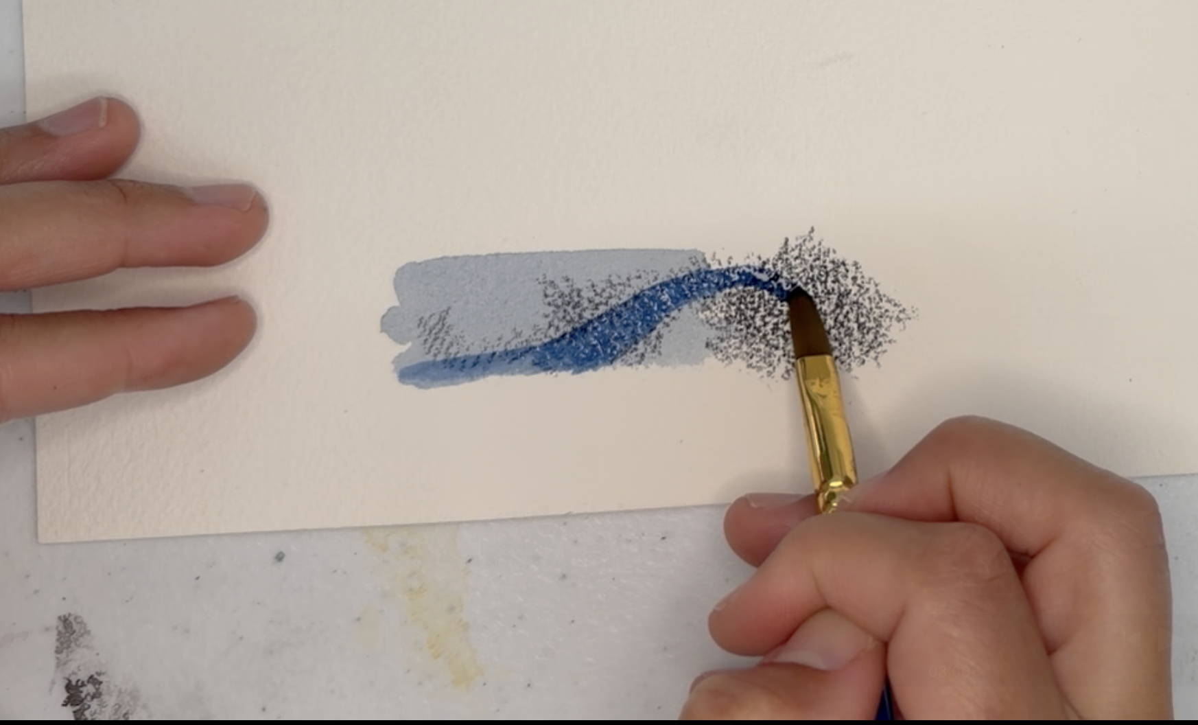 Photograph showing a close-up of hands painting on white paper with a flat brush, activating the dry Inktense material. The brush creates a textured effect with darker speckles concentrated at the right edge of the painted area.