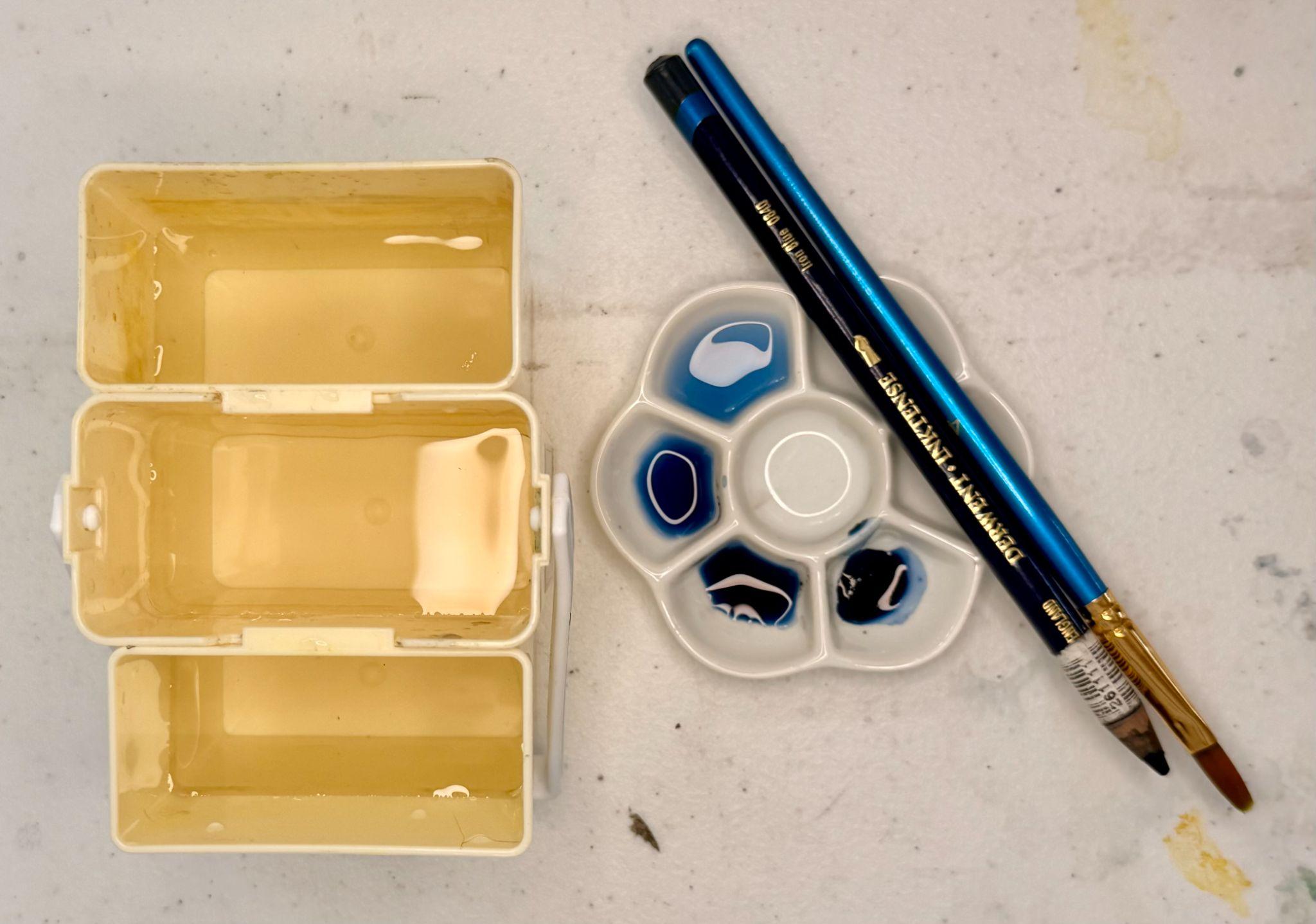 Photograph showing watercolor painting supplies arranged on a light surface, including a three-compartment water container, a small flower-shaped palette with blue paint, and two blue-handled paintbrushes. The setup highlights tools used for mixing and applying watercolor paints, with visible paint residue and water reflections indicating recent use.