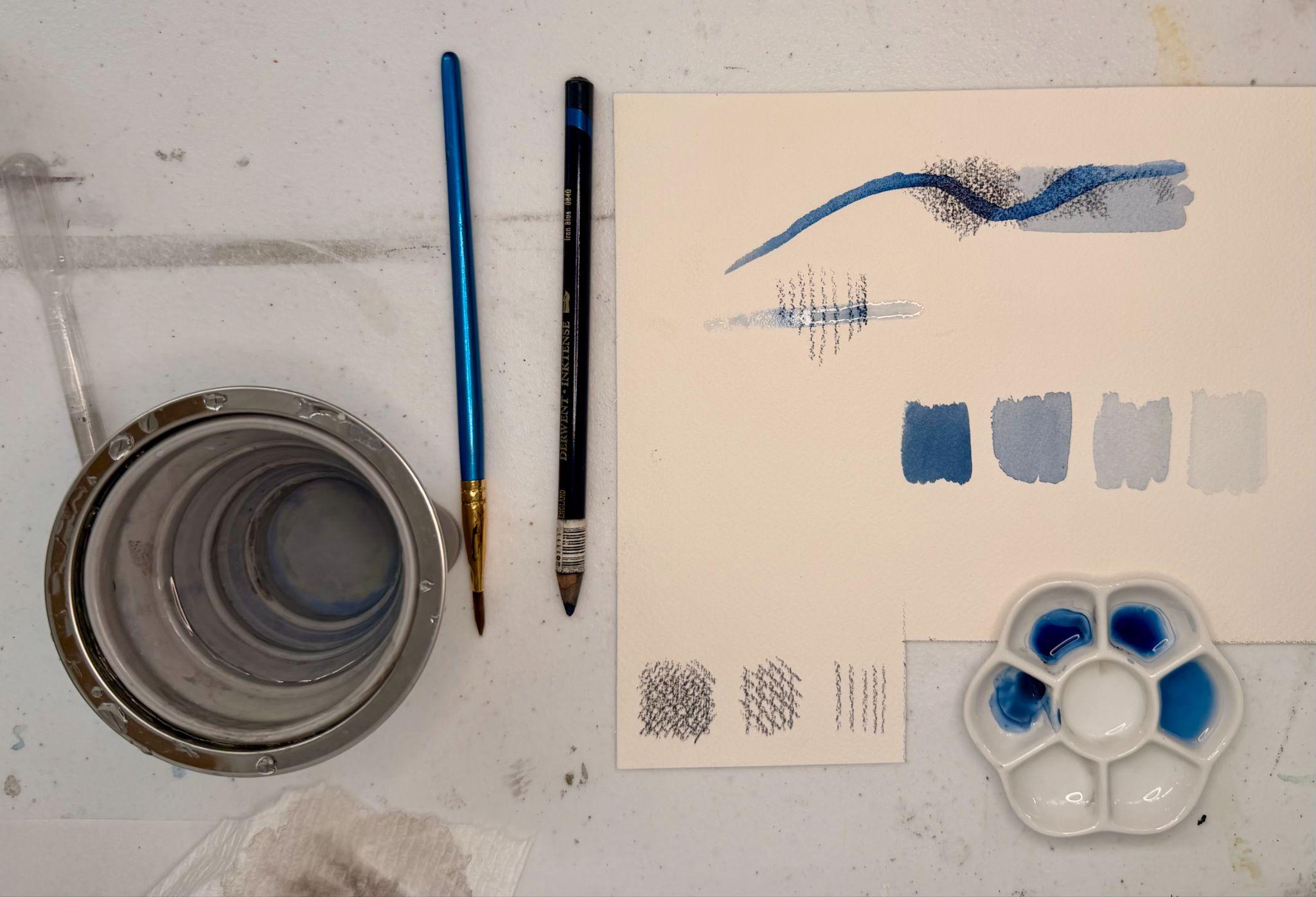 Photograph of a watercolor painting setup showing two paintbrushes, a metal cup with water, a palette with blue paint, and a paper with blue paint swatches and brushstroke tests. The paper displays gradients of blue shades, various brushstroke textures, and a curved brushstroke, highlighting experimentation with color intensity and brush techniques.