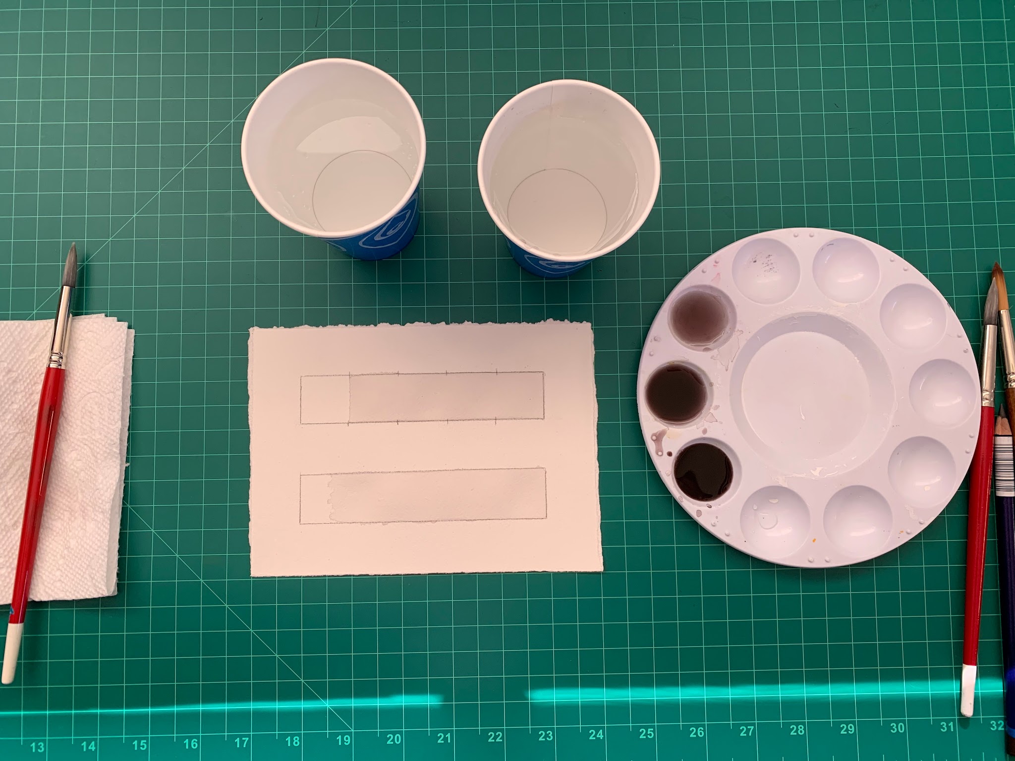 Photograph of a watercolor painting setup on a green cutting mat, featuring two paintbrushes, two paper cups with water, a white paint palette with brown and black paint, and a piece of paper with two rectangular outlines. The arrangement suggests preparation for a painting project focused on blending.