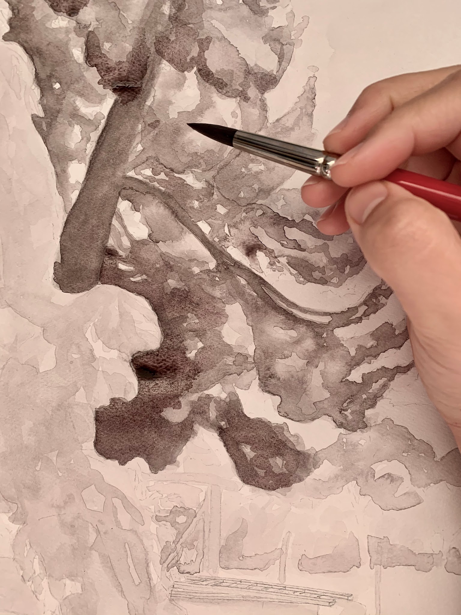 Photograph of a hand holding a paintbrush applying dark watercolor to a detailed leaf illustration. The artwork features varying shades of gray and black to create depth and texture in the leaf's veins and edges.