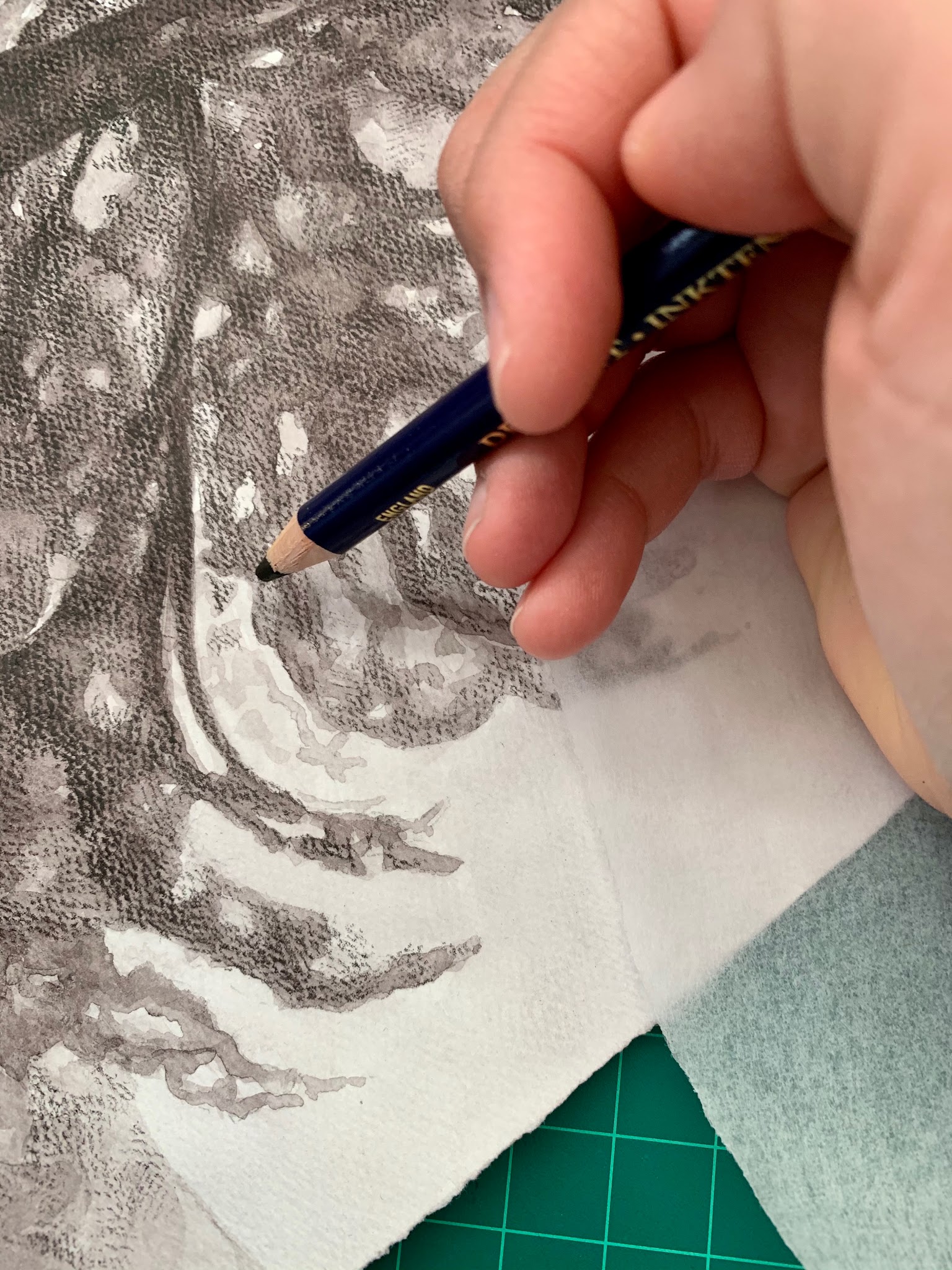 Photograph showing a close-up of a hand using a pencil to create detailed shading on a textured drawing. The artwork features intricate dark and light areas, with the hand positioned over a green cutting mat and a piece of translucent paper nearby.