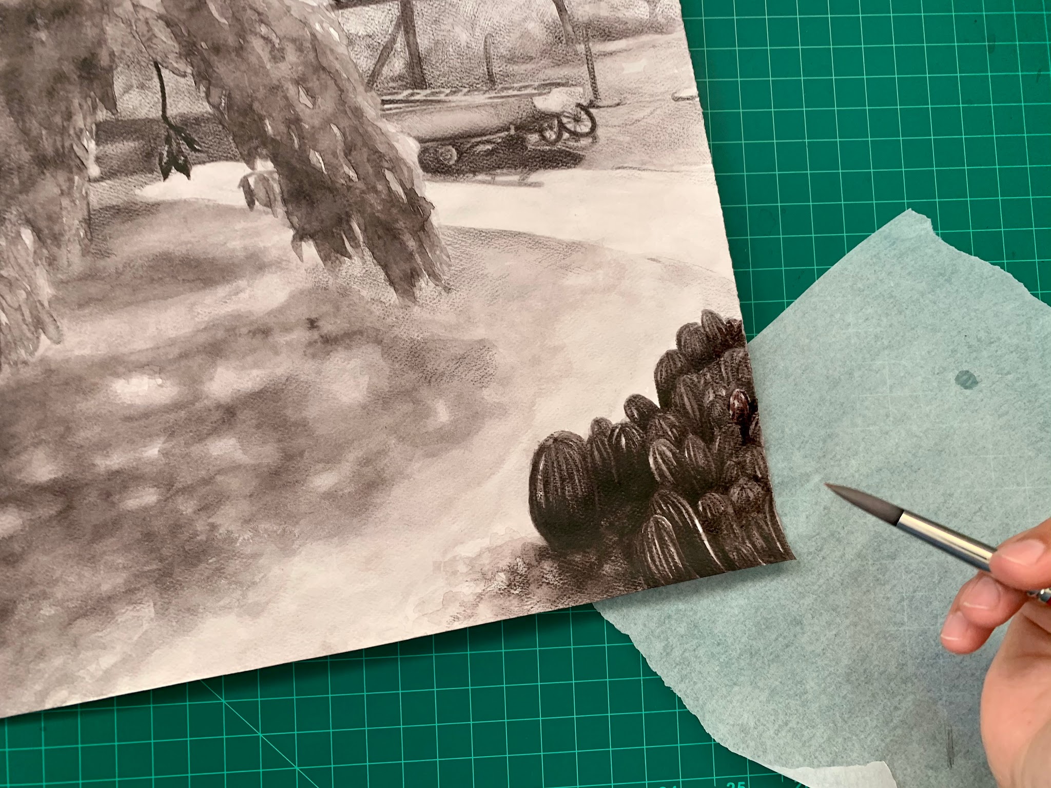 Photograph of a detailed black-and-white watercolor pencil drawing depicting a park scene with trees and a boat with fence in the background. A hand holding a paintbrush and a piece of tissue paper are visible on a green cutting mat, suggesting ongoing artwork refinement or shading.