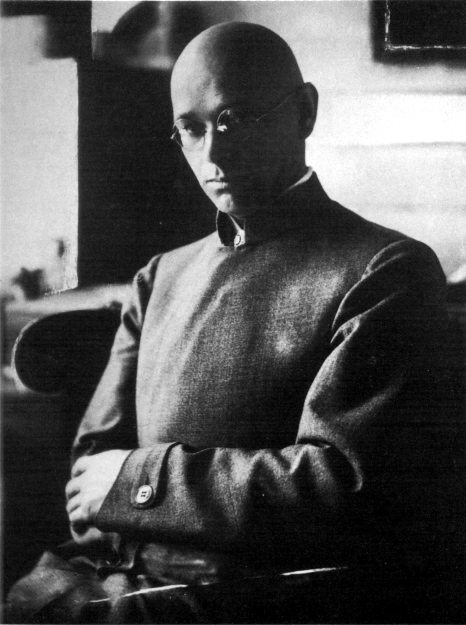 Black and white photograph of a man with a shaved head and round glasses sitting with arms crossed, wearing a high-collared jacket. The setting appears indoors with soft lighting highlighting the contemplative expression on the man's face.