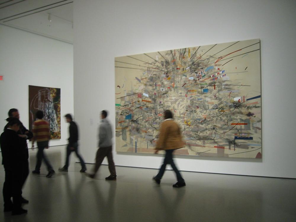 Photograph of an art gallery interior showing visitors walking past two large abstract paintings mounted on white walls. One painting features a chaotic explosion of lines and colors radiating from a central point, while the other displays darker, more concentrated abstract forms.