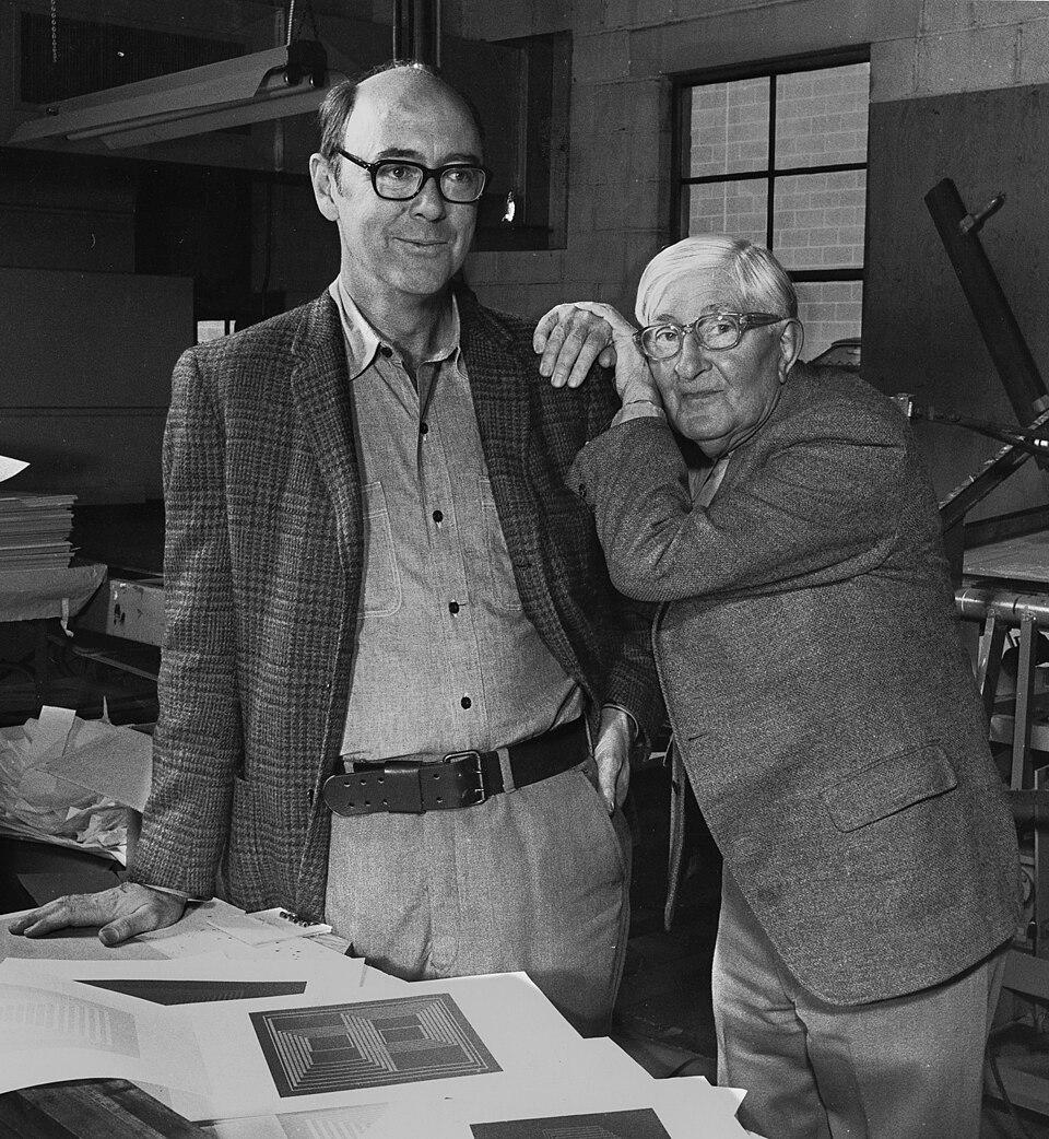 Black and white photograph of two older men standing in a workshop or studio, with one man leaning on the other's shoulder. The setting includes papers and architectural or design drawings on a table, suggesting a creative environment.