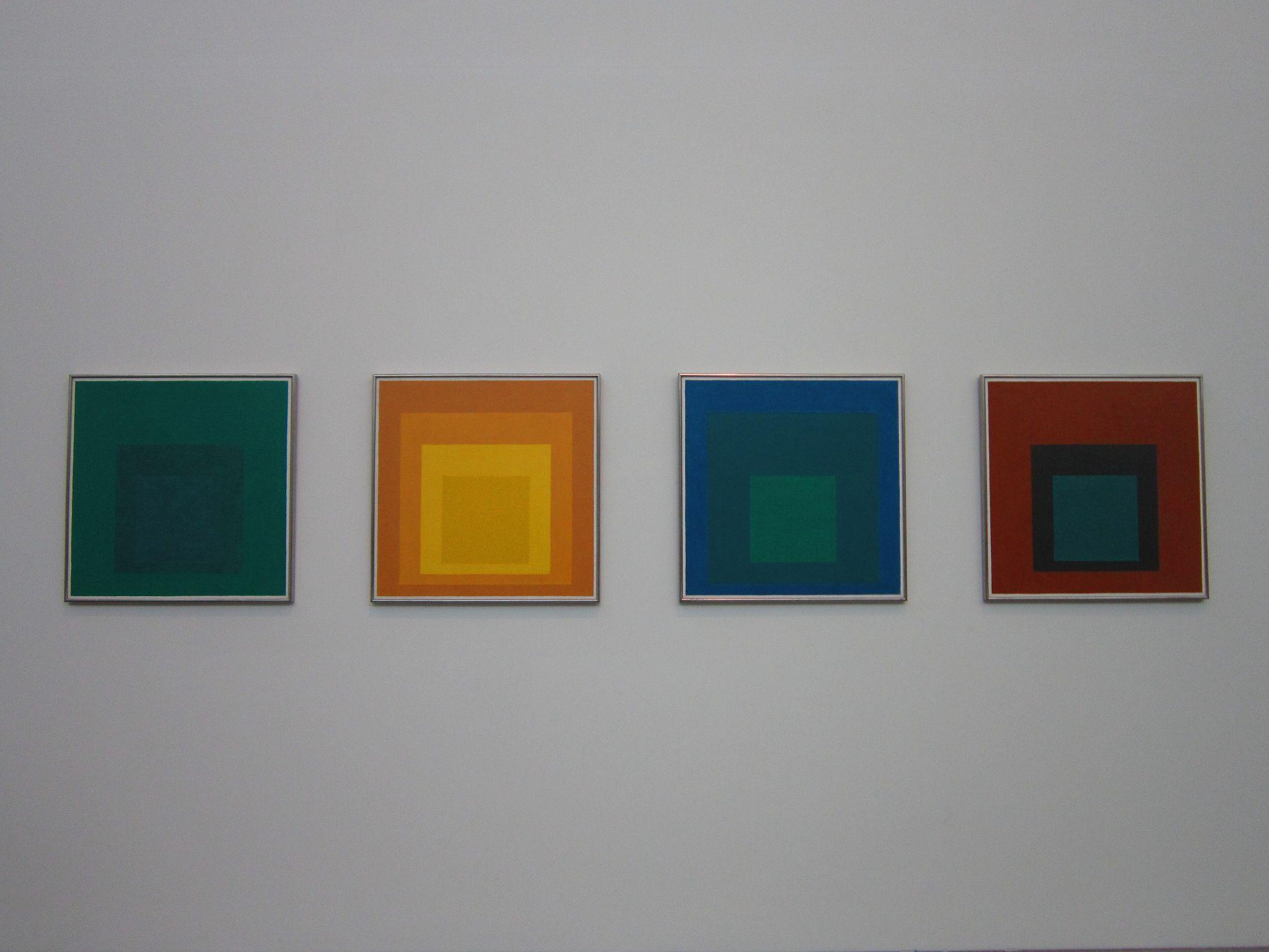 Photograph of four framed abstract artworks featuring nested squares in varying color schemes, displayed on a plain white wall. Each artwork uses contrasting shades—green, orange-yellow, blue-green, and red-teal—highlighting color interaction and geometric simplicity.