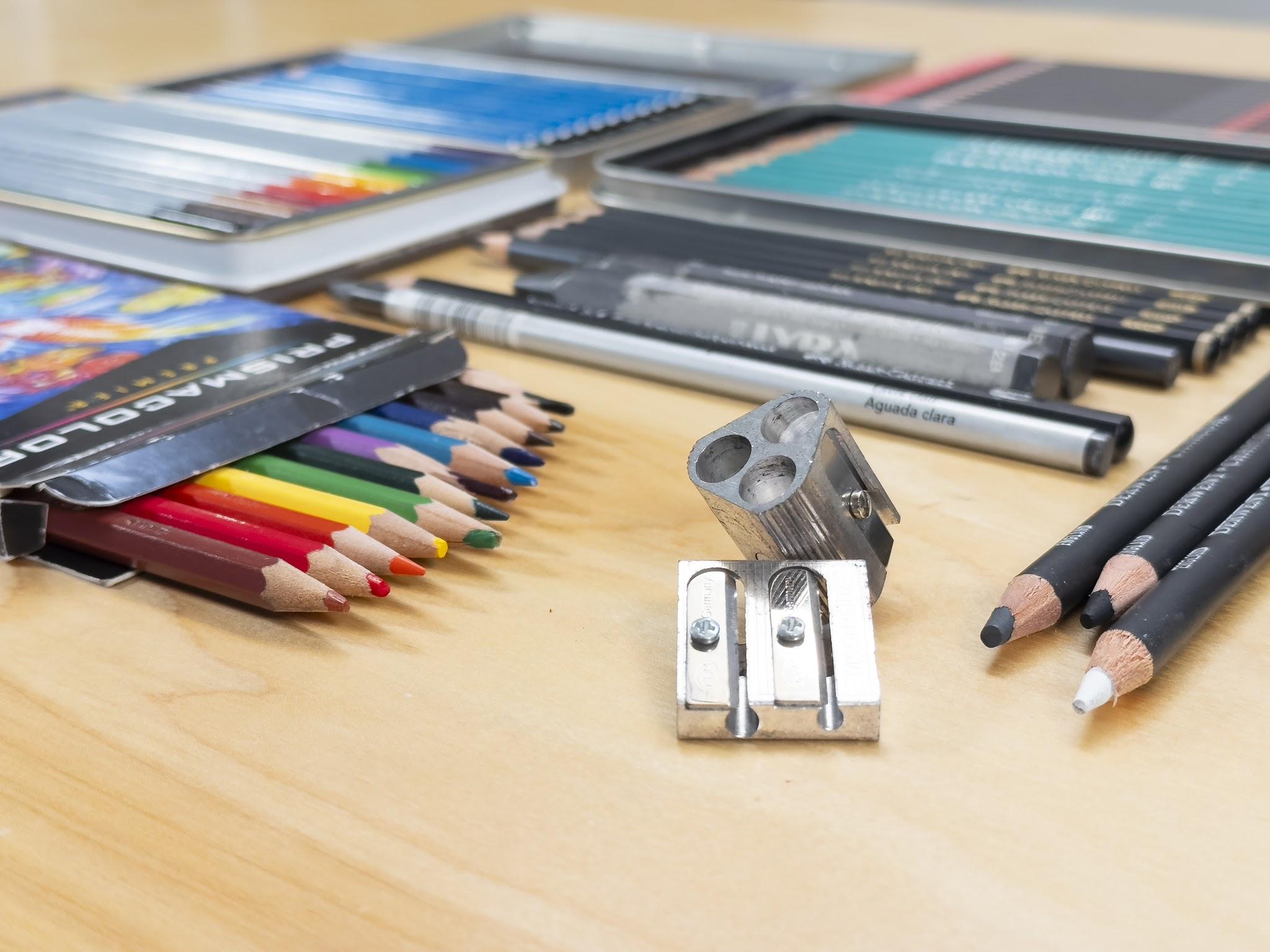 Photograph showing various art supplies arranged on a wooden surface, including colored pencils in a box, graphite pencils, charcoal sticks, and two metal pencil sharpeners. The image highlights a range of drawing tools with different colors, textures, and sizes, emphasizing preparation for artistic work.