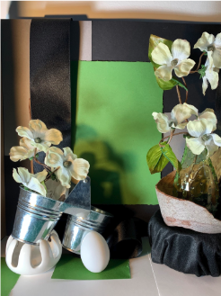 Photograph of a still life arrangement featuring white flowers in a glass vase and small metal buckets, set against a green and black background. The composition includes a white egg, a black ribbon, and a white geometric object, creating a contrast of textures and colors.