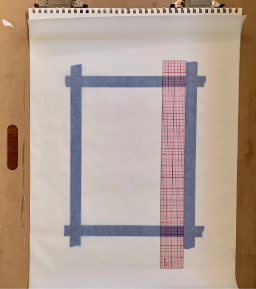 A photograph showing a blank sketchpad page taped to a wooden surface with blue painter's tape forming a rectangular border. A transparent ruler with red grid lines is placed vertically along the right side of the taped area, highlighting measurement and alignment.