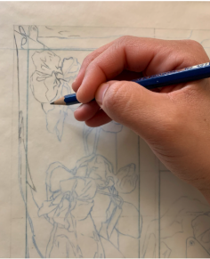 Photograph of a hand holding a blue pencil sketching floral designs on paper. The drawing features detailed outlines of flowers and leaves within a rectangular border, highlighting the artist's focus on intricate botanical elements.