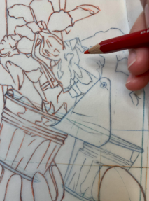 Photograph of a hand using a red colored pencil to trace or color a detailed line drawing featuring a flower pot and a birdhouse. The drawing includes brown and blue outlines with intricate floral and structural elements.