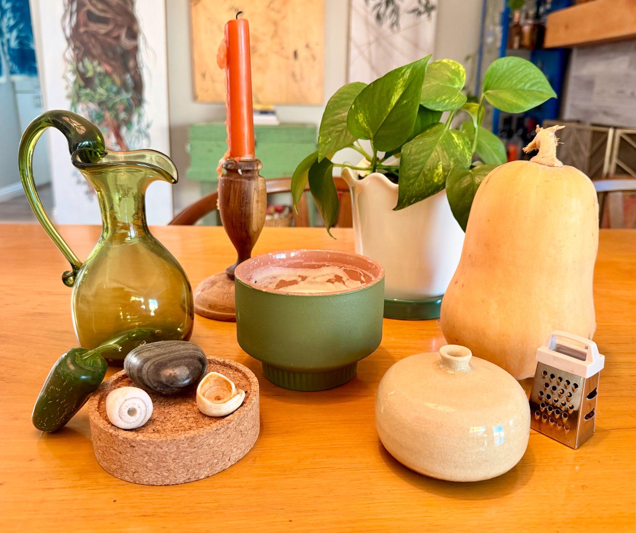 Photograph of a tabletop arrangement featuring a green glass pitcher, an orange candle in a wooden holder, a green cup, a potted plant in a white vase, a butternut squash, a small ceramic container, a mini grater, and a cork coaster holding a jalapeño and three seashells. Warm lighting highlights natural textures and earthy tones, creating a cozy and eclectic still life composition.