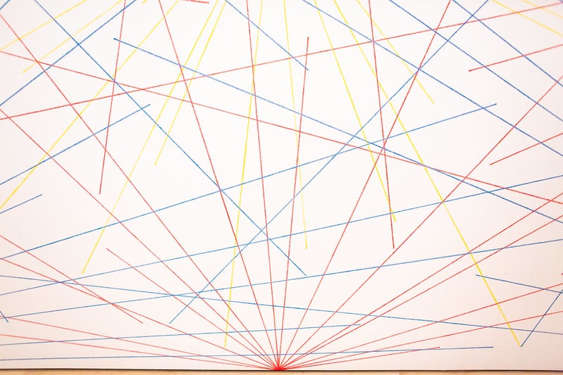 Abstract photograph featuring numerous intersecting straight lines in red, blue, and yellow on a white background. Lines radiate from a single point at the bottom center, creating a geometric pattern with varying angles and lengths.