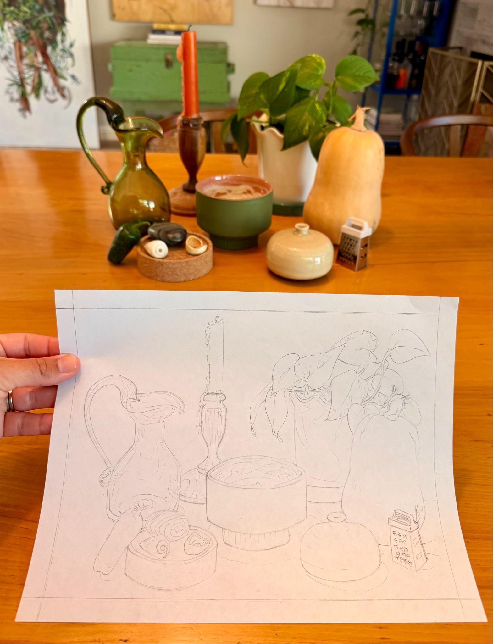 Photograph of a hand holding a pencil sketch replicating a still life arrangement on a wooden table, featuring a green pitcher, orange candle, potted plant, butternut squash, small grater, and various containers. Sketch outlines key objects and composition, capturing shapes and placement while omitting colors and fine details present in the original scene.