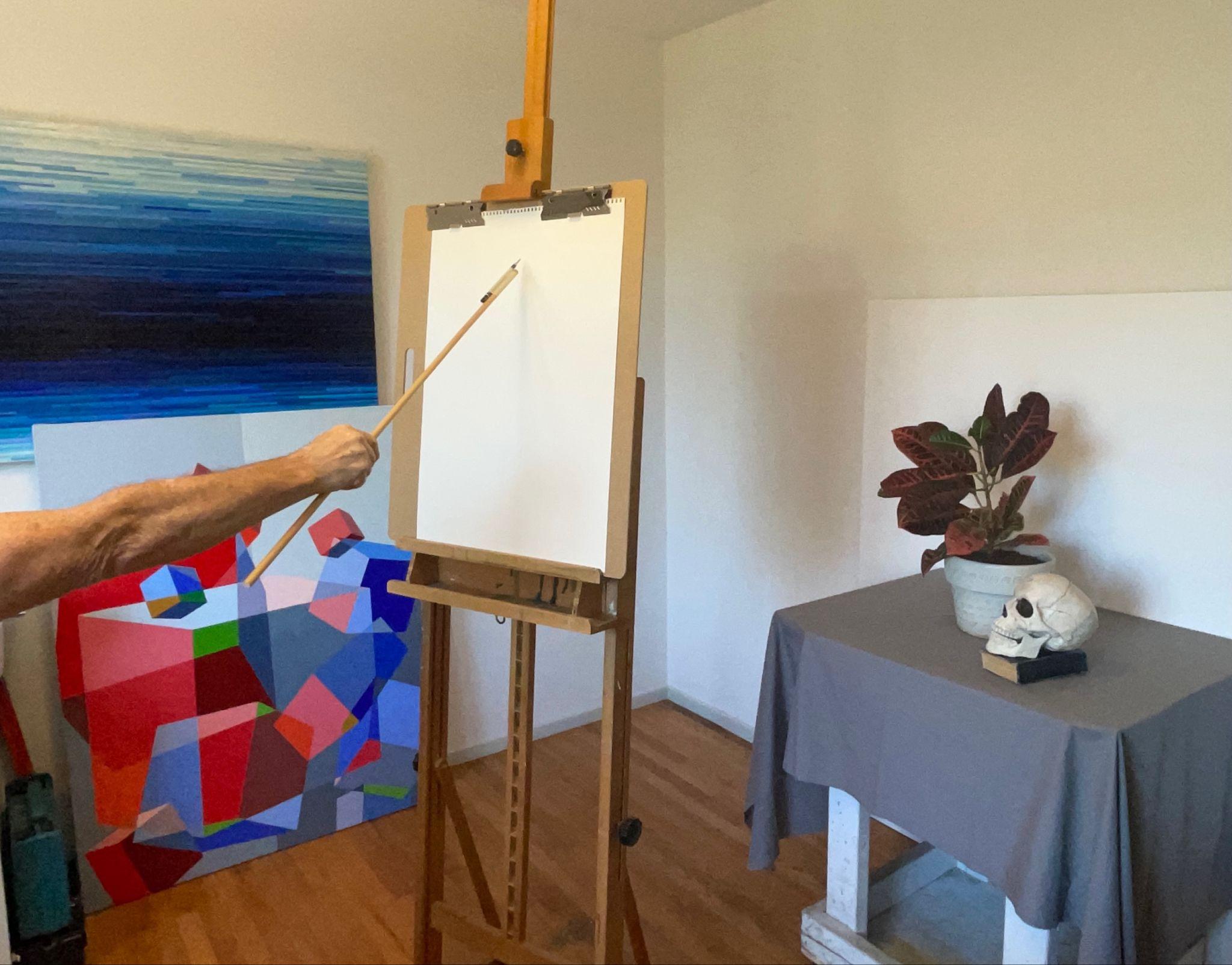 Photograph of an artist's studio featuring a wooden easel with a blank canvas and a hand holding a wooden dowel with a pencil attached directed at the canvas. The room includes colorful abstract paintings leaning against a wall and a table covered with a gray cloth displaying a potted plant and a human skull model.