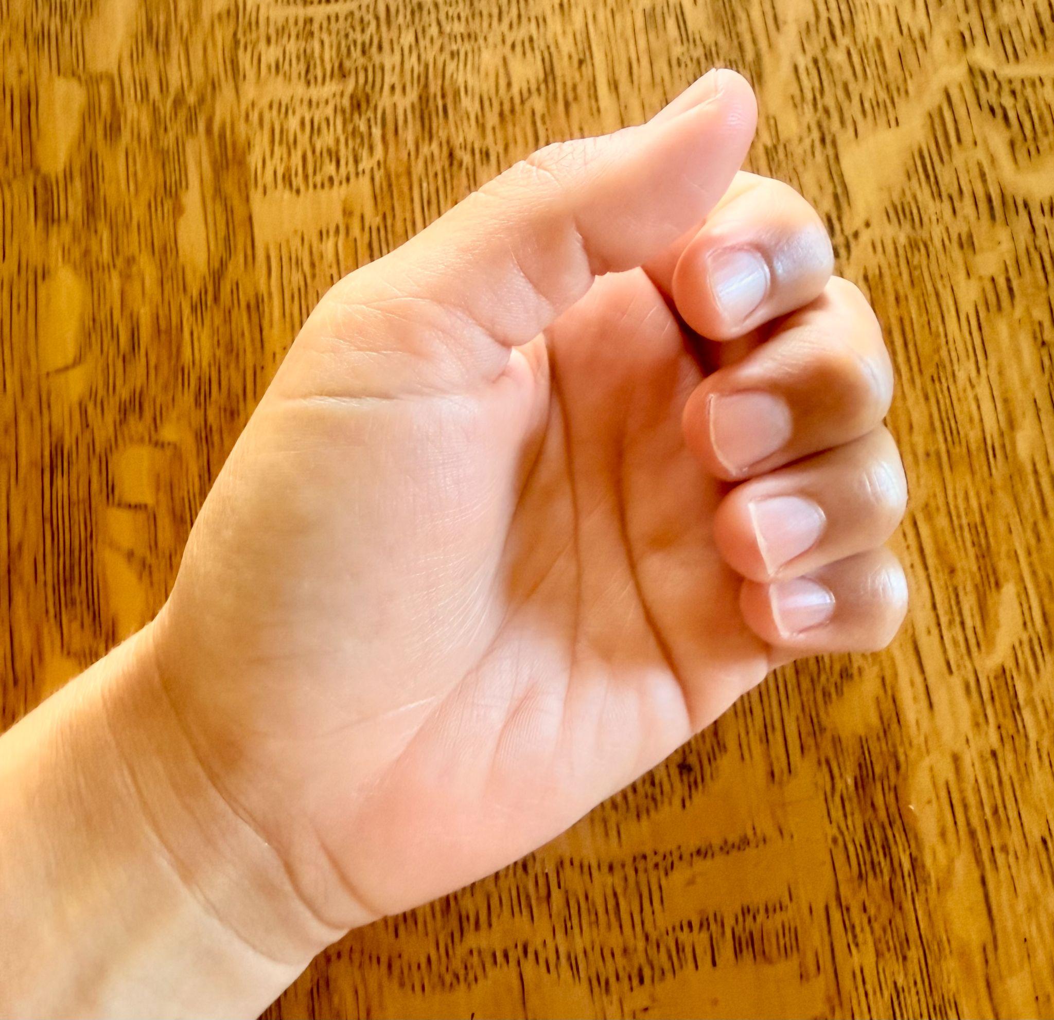 Photograph of a left hand with fingers curled inward against a wooden surface background. The hand shows natural skin tone and short, clean fingernails, with visible palm lines and slight shadows.