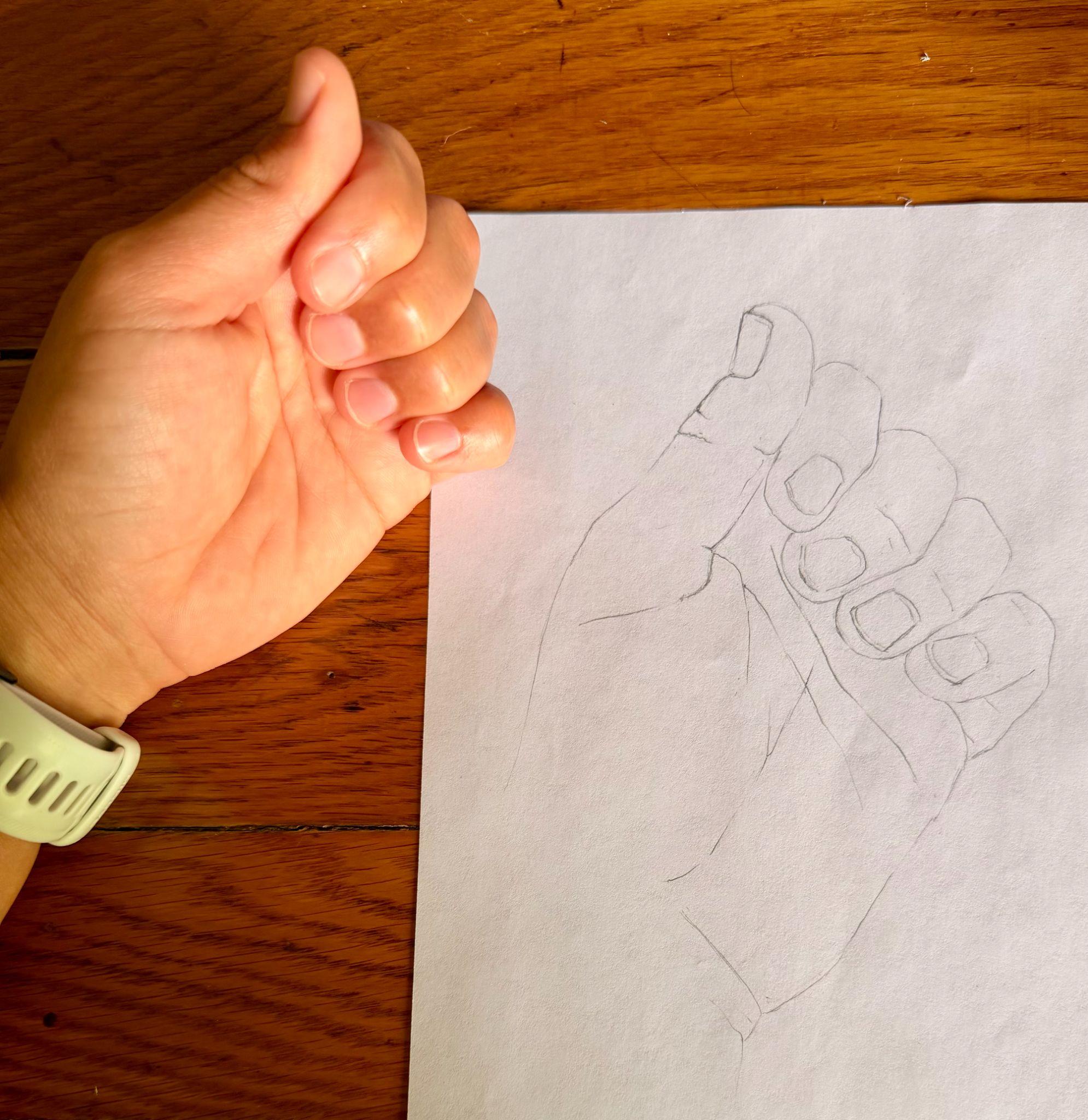Photograph showing a left hand with fingers curled into a loose fist next to a pencil sketch of the same hand on white paper. The hand wears a light-colored wristband, and the sketch outlines finger shapes and nails with simple lines on a wooden surface background.