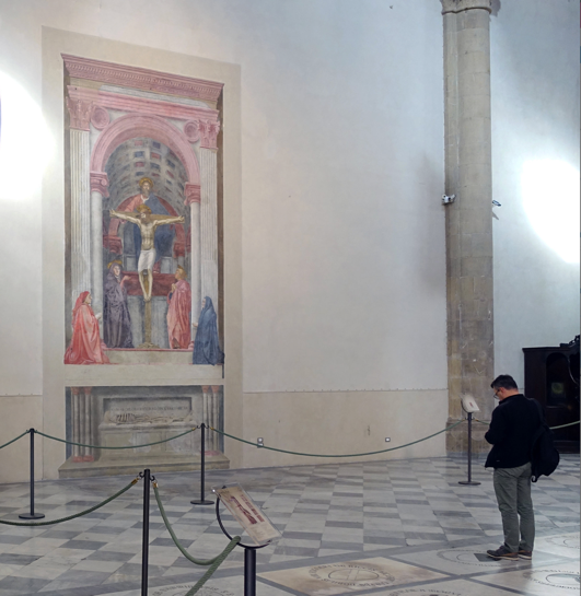 Photograph of a large religious fresco inside a church, depicting a crucifixion scene with figures in red, purple, and blue robes surrounding the cross under an architectural arch. The fresco is roped off for protection, with a visitor standing nearby on a checkered marble floor, observing the artwork.
