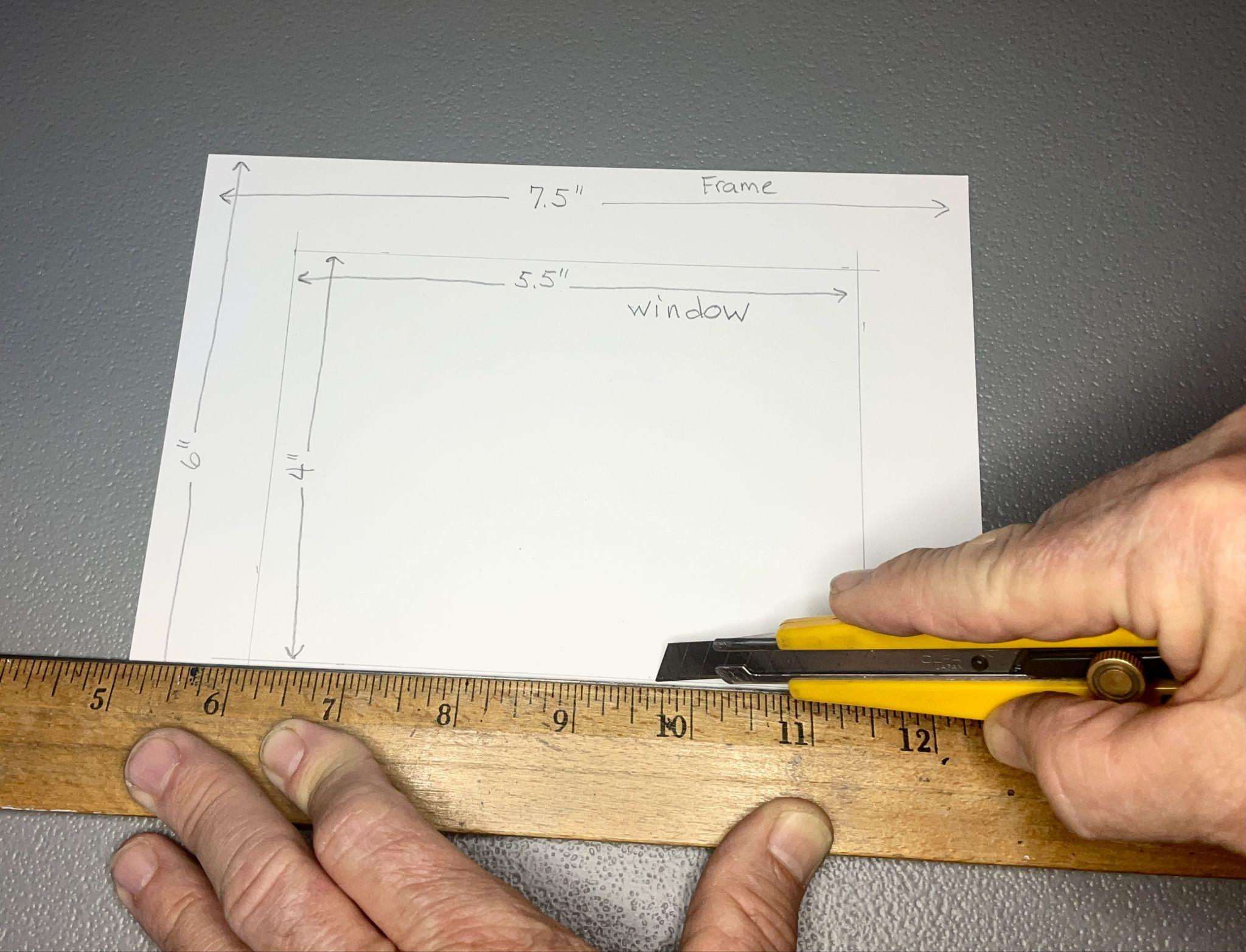 Photograph showing hands measuring and cutting a white paper viewfinder with a utility knife and wooden ruler on a gray surface. Paper contains hand-drawn rectangular frame and window outlines labeled with dimensions 7.5" by 6" for frame and 5.5" by 4" for window.