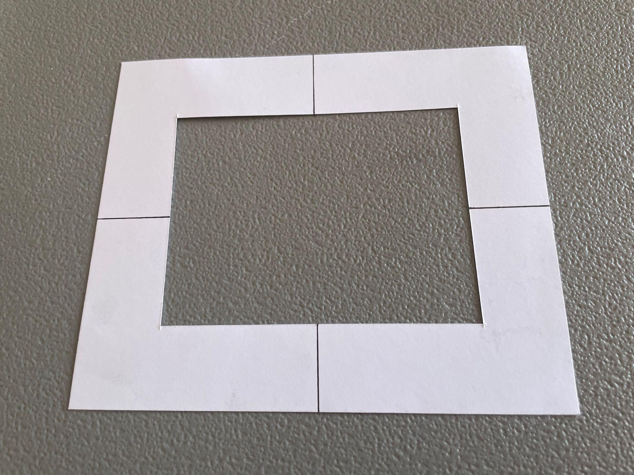 Photograph of a white paper viewfinder frame with a rectangular cutout in the center, placed on a textured gray surface. Frame edges are marked with four black lines dividing each side into two segments, highlighting the rectangular shape.