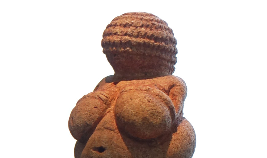 Photograph of a detail of Venus of Willendorf, a small Paleolithic figurine representing a female figure with exaggerated breasts. The sculpture features detailed braided hair and is believed to symbolize fertility.