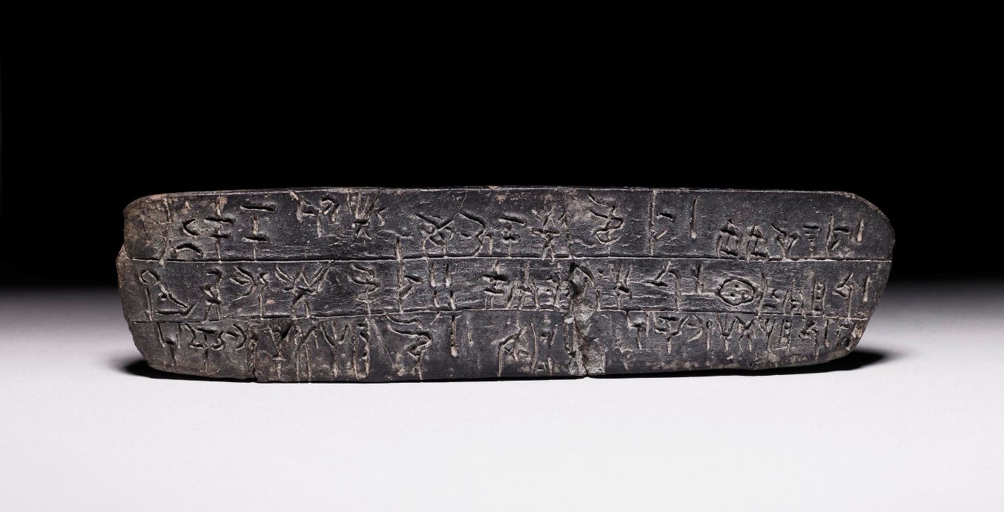 Photograph of a stone tablet inscribed with Linear B script arranged in horizontal rows. The dark tablet contrasts with a light surface and black background, highlighting detailed carvings and signs of wear on the artifact.