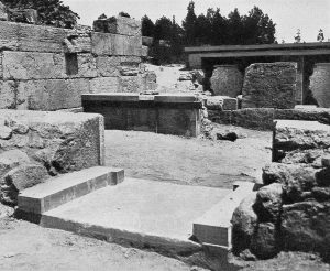 Black and white photograph of ancient stone ruins featuring rectangular platforms, steps, and large stone blocks arranged in an open area. Background includes trees and a partially visible stone structure, highlighting archaeological site details and historical architecture.