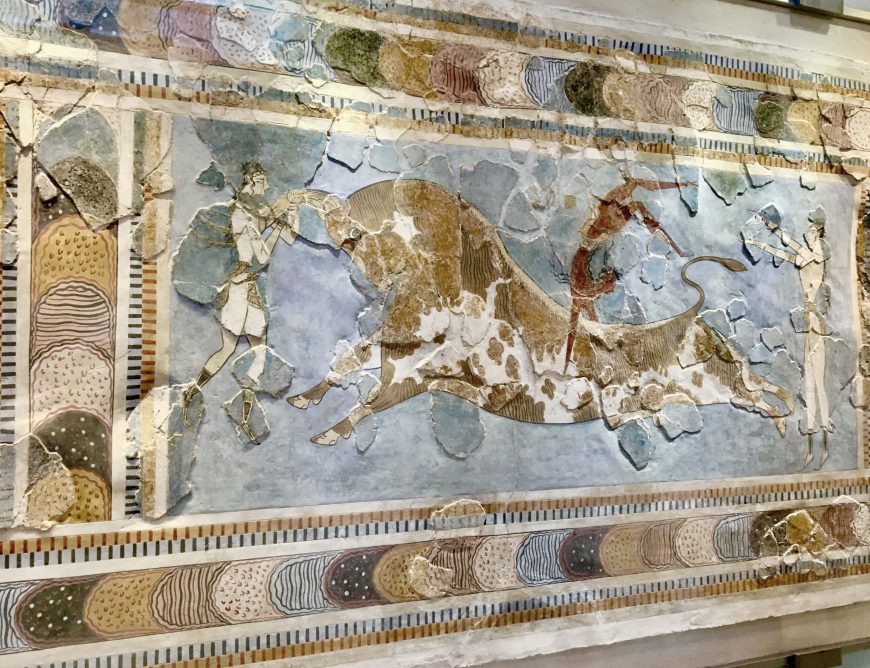 A fresco painting depicting a bull-leaping scene from ancient Minoan culture, showing three human figures interacting with a large bull in dynamic motion. The artwork features earthy tones with detailed borders of patterned semicircles and stripes, highlighting cultural and ritualistic significance.