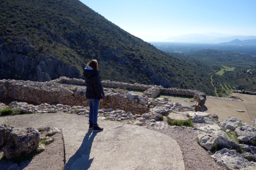 Photograph of a person standing on ancient stone ruins overlooking a vast valley with mountains and sea in the distance. The scene highlights historical architecture with rugged stone walls and natural landscape under clear blue sky.