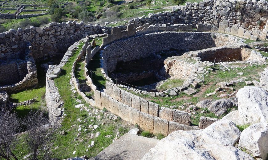 Photograph of the “Palace” and Grave Circle A featuring a circular structure with tiered seating resembling an amphitheater or ceremonial site. The scene highlights weathered stone walls, grassy patches between tiers, and surrounding rocky terrain.