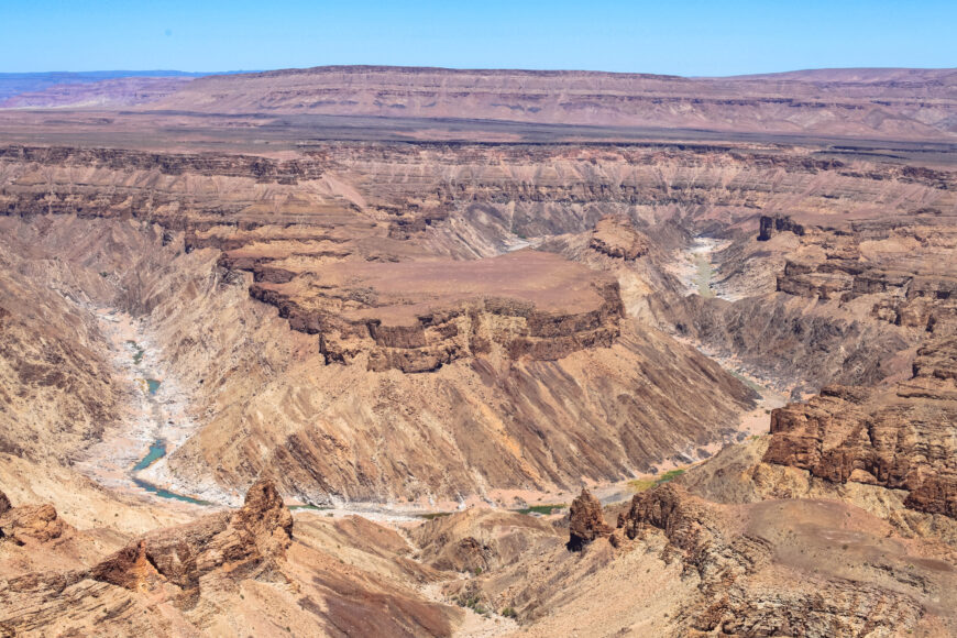Photograph of a vast canyon landscape featuring steep, layered rock formations and a winding river at the canyon floor. The scene highlights geological strata with varying shades of brown and tan under a clear blue sky, emphasizing erosion patterns and natural terrain contours.