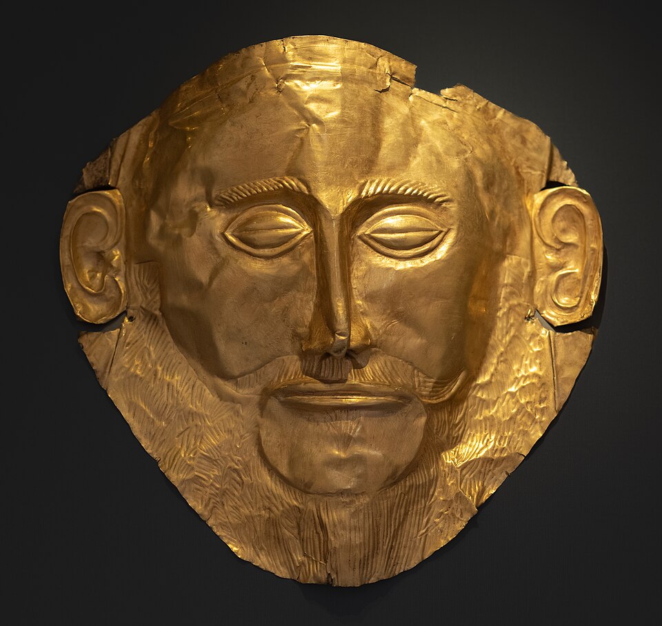 Photograph of a gold funerary mask shaped like a human face with detailed eyes, nose, mouth, ears, and beard. The mask features a hammered texture and is displayed against a dark background, highlighting its metallic sheen and craftsmanship.