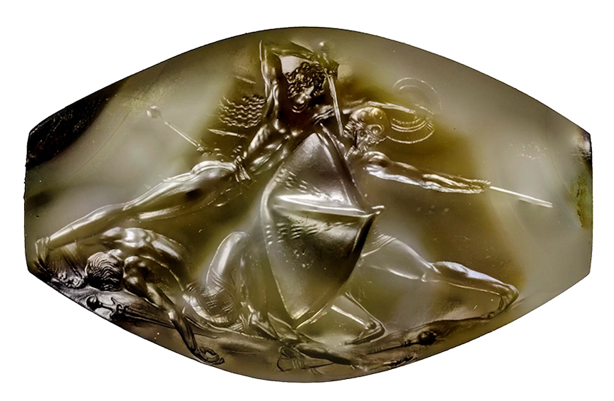 Photograph of an ancient carved gemstone depicting three warriors in dynamic battle poses, with one holding a shield and spear and another wielding a sword. The translucent stone features intricate details highlighting muscular forms and flowing hair, emphasizing movement and conflict.
