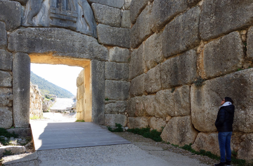 Photograph of Lion Gate, Mycenae with massive, weathered blocks forming walls and a lintel featuring carved reliefs. A person stands near the right wall, highlighting scale, while sunlight illuminates a pathway leading through the gateway toward distant hills.