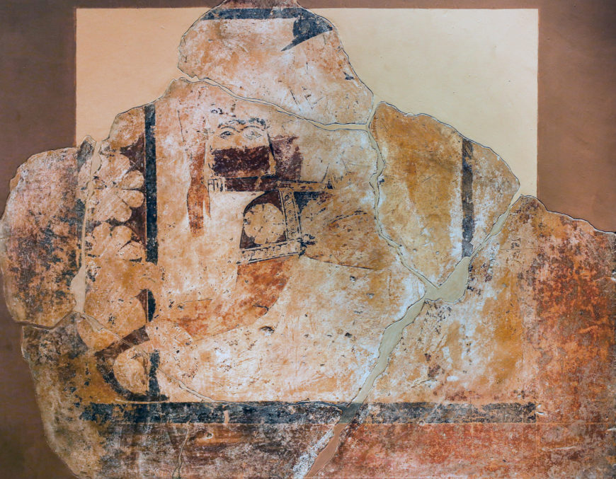 Fresco fragment depicting a partially preserved figure with geometric and floral patterns, showcasing ancient artistic techniques. Colors include faded reds, blacks, and browns, with visible cracks and missing sections highlighting age and deterioration.
