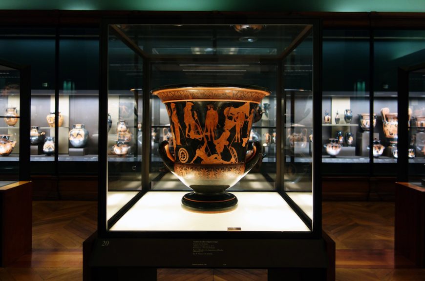 Photograph of an ancient Greek terracotta vase displayed in a glass case within a museum gallery. Vase features red-figure artwork depicting human and mythological figures, surrounded by other similar pottery pieces on illuminated shelves in the background.