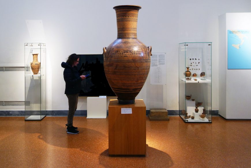 Photograph of a museum exhibit featuring the Dipylon Amphora displayed on a wooden pedestal, surrounded by glass cases containing smaller pottery pieces. A visitor stands nearby, looking at a phone, with informational placards and a map visible on walls in the background.