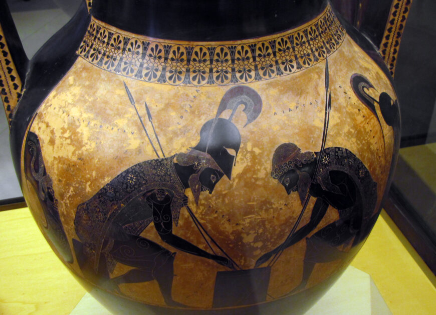 Photograph of an ancient Greek black-figure pottery vase depicting two armored warriors crouching and playing a board game, representing a scene from the Trojan War. The vase features intricate detailing with black figures against a reddish-brown background, bordered by a decorative floral pattern near the top.